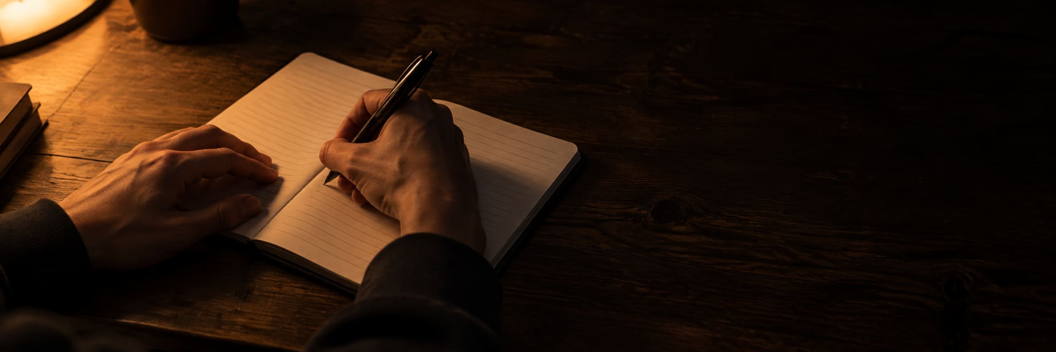 Two hands writing in a notebook on a dark wooden table, lit by a warm lamp, with a mug and two books beside them.