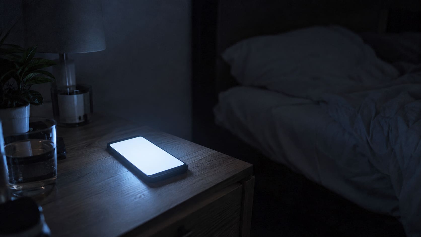 A dark bedroom at night. A phone lies face up on the nightstand, its bright screen the only light in the room.